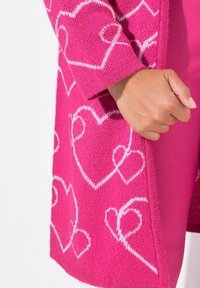 Růžový pletený svetřík se vzorem bílých srdcí, volného střihu a žebrovanými manžetami. Materiál vypadá měkký a strukturovaný.