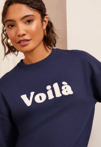 Merinovärisen hupparin edessä on fluffy-valkoisella kirjailtuna sana "Voilà". Hupparissa on ribbaus kaula-aukossa ja pehmeä pinta, esitettynä neutraalilla taustalla.