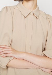 Beige blus med krage, plisserad front och puffärmar. Mjuk, texturerad tyg med en enda knappdetalj vid kragen.