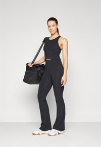 Black athletic crop top and flared pants made from stretchy fabric. Model carries a black duffel bag with mesh pocket. White sneakers complete the look.