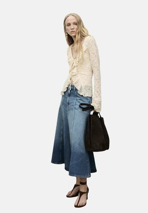 Haut en dentelle crème avec des volants, associé à une jupe en denim à jambes larges déclinant du bleu foncé au bleu clair. La mannequin tient un sac marron foncé et porte des sandales noires.