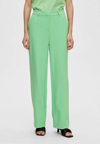 Pantaloni larghi di un verde chiaro con una texture liscia, vita alta e passanti per cintura, abbinati a sandali neri con punta aperta.