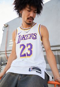 Bijela košarkaška majica s ljubičastim i žutim detaljima, koja sadrži natpis "LAKERS" i broj "23", izrađena od mrežaste tkanine, označena nba i Nike brendiranjem.