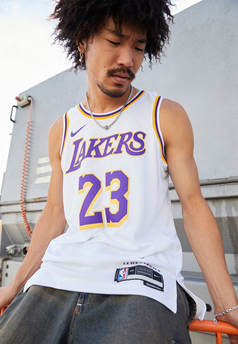 Bijela košarkaška majica s ljubičastim i žutim detaljima, koja sadrži natpis "LAKERS" i broj "23", izrađena od mrežaste tkanine, označena nba i Nike brendiranjem.
