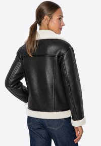 Czarna kurtka z sztucznej skóry z białym kołnierzem i mankietami z owczej wełny, charakteryzująca się gładką fakturą i skróconym krojem. Pokazany widok z tyłu.