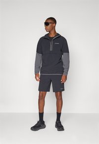 Čierna a sivá zipová bunda s vysokým golierom, kombinovaná s čiernymi šortkami. Oblečenie je vyrobené z ľahkého materiálu s akcentmi loga.