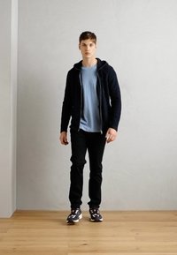 Felpa nera con zip sopra una maglietta azzurra, abbinata a pantaloni neri e sneakers grigie. Sfondo in legno, parete semplice.