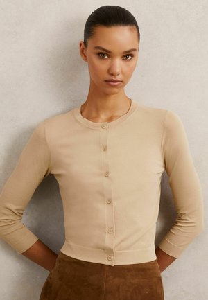 Frau mit zurückgestrichenen Haaren, die eine beige geknöpfte Strickjacke und einen braunen Wildlederrock trägt und vor einer schlichten hellen Wand steht.
