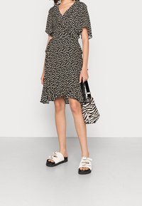 Robe noire avec motif de feuilles blanches, jupe évasée et manches courtes. Sandales blanches avec des lanières et semelles noires. Sac à main imprimé zèbre.