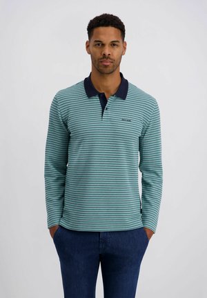 Mann mit kurzen lockigen Haaren, der ein langärmeliges Polohemd mit hellgrünen und marineblauen Streifen sowie dunkelblaue Hosen trägt, die Hände in den Taschen, vor einfarbigem Hintergrund stehend.