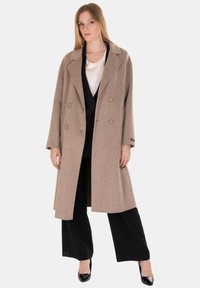 Cappotto doppiopetto in lana color marrone chiaro, con ampi risvolti, vestibilità oversize, maniche lunghe e taglio dritto. Abbinato a un completo nero.