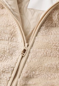 Gros plan d'une veste polaire beige avec une fermeture éclair en plastique visible partiellement ouverte, montrant une doublure intérieure lisse et un tissu extérieur doux et texturé.