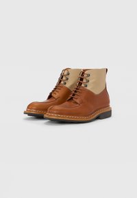 Bruine leren laarzen met een beige stoffen bovenkant, voorzien van ronde veters, metalen oogjes en een gestructureerde zool. Contrasterende stiksels langs de rand.