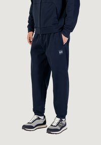 Dunkelblauer Fleece-Trainingsanzug mit einer Reißverschluss-Hoodie und elastischen Hosen, die mit einem Logo-Patch versehen sind. Die Schuhe sind grau und navyfarben.