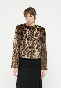 Joven vistiendo una chaqueta de piel de leopardo marrón y beige con grandes botones negros y una falda negra, de pie contra un fondo blanco.