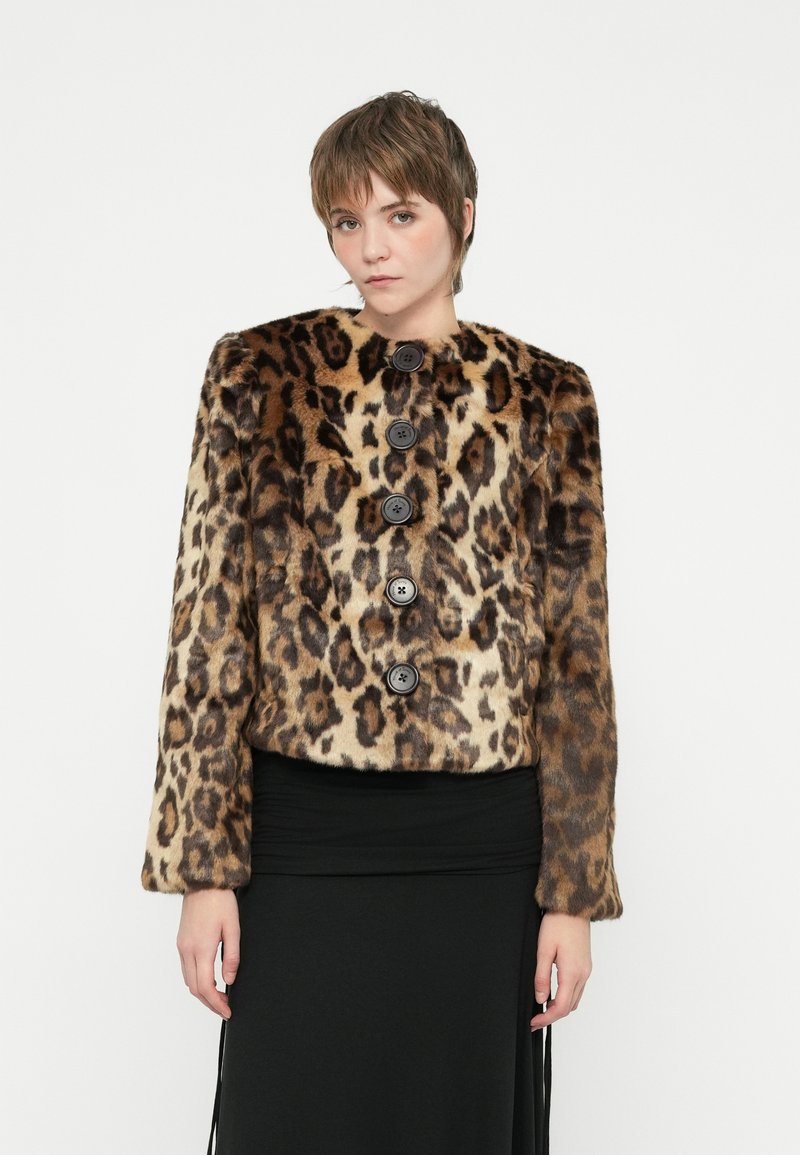 Ung person iført en brun og beige leopardmønstret pelsjakke med store sorte knapper og en sort nederdel, stående mod en hvid baggrund.