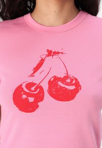 Nära bild av en rosa skjorta med en röd grafisk design av två sammanlänkade körsbär på framsidan, bärs av en person.