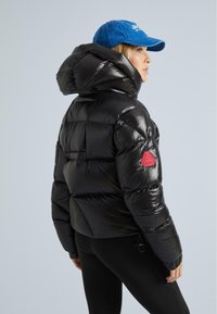 Donna che indossa una giacca a vento nera lucida con cappuccio e un berretto blu, mostrata di profilo su uno sfondo chiaro e semplice.