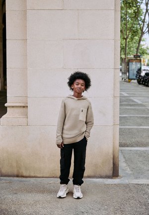 Ragazzo con capelli ricci in felpa beige con cappuccio, pantaloni cargo neri e scarpe da ginnastica bianche in piedi contro un muro di pietra su un marciapiede urbano.