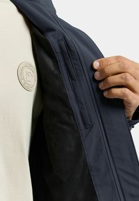 Marineblauw jack met een zachte binnenvoering, met een horizontale ritssluiting en een beige stoffen overhemd met een ronde logo.