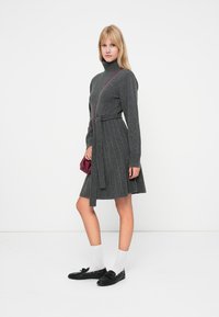 Šedé pletené šaty s rolákem, těsným živůtkem a rozšířenou sukní. Modelka drží malou burgundskou tašku; má na sobě černé boty a bílé ponožky.
