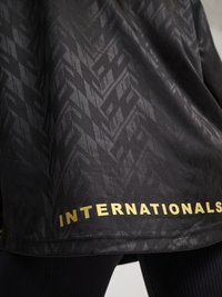 Svart stoff med subtilt geometrisk mønster og gulltekst «INTERNATIONALS» nær kanten, båret over svarte ribbestrikkede leggings.