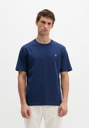 Jonge man met kort bruin haar die een effen donkerblauw T-shirt en lichtbeige broek draagt, staand tegen een witte achtergrond.