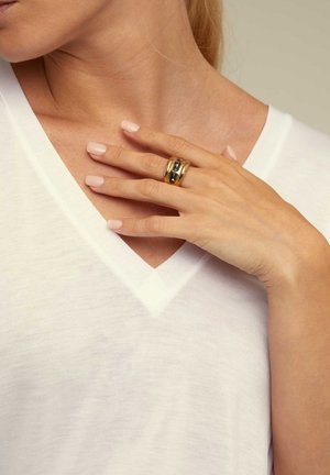 Mujer vistiendo una blusa blanca de cuello en V, con la mano de uñas nude pulidas descansando sobre el pecho, mostrando un anillo dorado ancho con piedras oscuras.