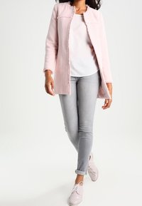 Manteau texturé rose pâle, à coupe droite, ouvert sur le devant et sans fermeture, porté sur un haut blanc et un jean gris. Accentué par des chaussures claires.