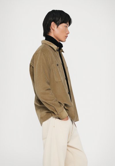 Camicia in velluto a coste marrone con colletto, due tasche sul petto e chiusura con bottoni, indossata sopra un dolcevita nero e abbinata a pantaloni chiari.