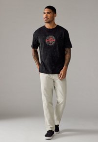 Zwarte katoenen t-shirt met een cirkelvormige rode grafische tekst "DE VERRADERS" in het midden, gecombineerd met lichtbeige broeken en zwarte sneakers.