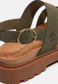 Sandale en cuir vert olive avec boucle en laiton réglable, semelle intérieure rembourrée tan embossée du logo Timberland, et semelle extérieure épaisse en caoutchouc marron.