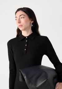 Zwarte geribbelde jurk met een kraag, voorzien van gouden knoopaccenten. Houdt een zwarte leren tas met een gladde textuur vast.