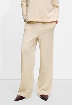 Pantaloni beige a gamba larga realizzati in un tessuto morbido, caratterizzati da una vestibilità rilassata e linee pulite. Abbinati a scarpe bordeaux a punta.