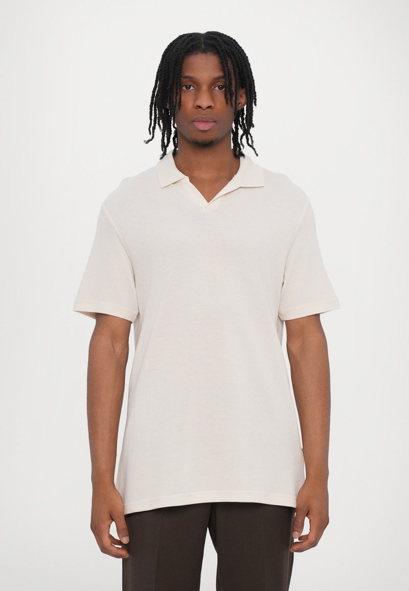 Polo léger beige clair à manches courtes avec un col en V, un col côtelé et une coupe décontractée. Texture douce et détails de design minimalistes.