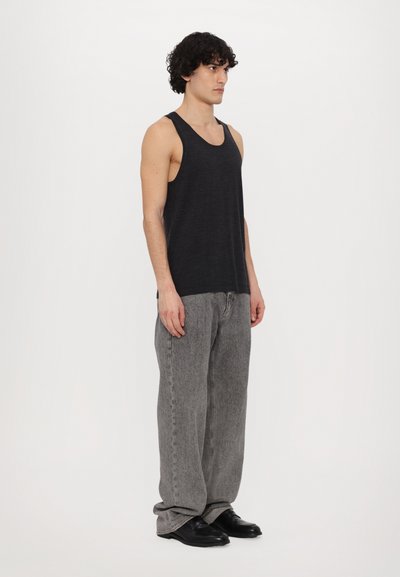 Débardeur noir avec des emmanchures larges et une texture douce, associé à un jeans gris clair ample et des chaussures noires, vu de côté.