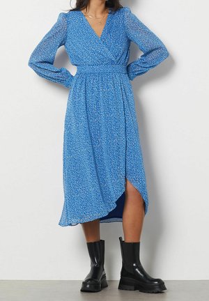 Kobieta w niebieskiej długiej sukience midi w kwiaty z dekoltem w serek i czarnych masywnych botkach, stojąca na tle jasnej ściany.