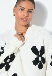 Weiß gestrickter Cardigan mit schwarzen Blumenmustern. Verfügt über einen V-Ausschnitt, lange Ärmel und gerippte Bündchen. Die Textur wirkt weich und gemütlich.