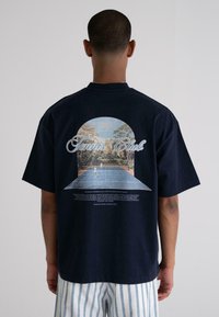 Navy blue oversized cotton t-shirt featuring a graphic print of a tennis court within an arched frame and text detailing a tennis club.