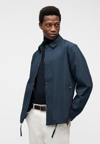 Άνδρας με υφασμάτινο μπλε σακάκι πάνω από μαύρο ζιβάγκο, με λευκό παντελόνι και καφέ ζώνη, στέκεται με τα χέρια στις τσέπες, κοιτώντας δεξιά.