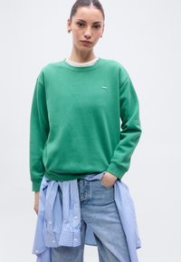 Sweat-shirt vert à col rond et poignets côtelés, superposé sur une chemise blanche. Chemise à rayures bleues et blanches, boutonnée et nouée à la taille.