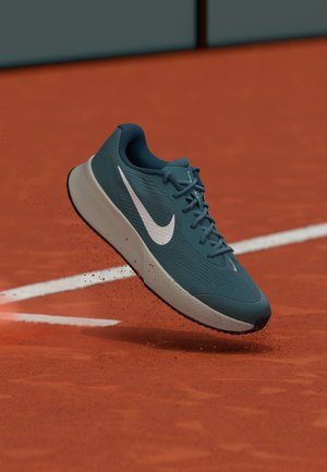 Sötét türkizkék Nike futócipő fehér Swoosh logóval a levegőben, egy salakos teniszpálya felett, rajta szétszórt salakrészecskékkel.