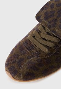 Sneaker z teksturowanym oliwkowo-zielonym i brązowym wzorem w panterę, z brązowymi sznurówkami oraz zaokrąglonym czubkiem, na beżowej gumowej podeszwie.