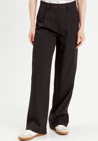 Pantalons larges noirs avec des plis devant, ceinture avec une fermeture à bouton et une texture lisse. Associés à des baskets blanches.