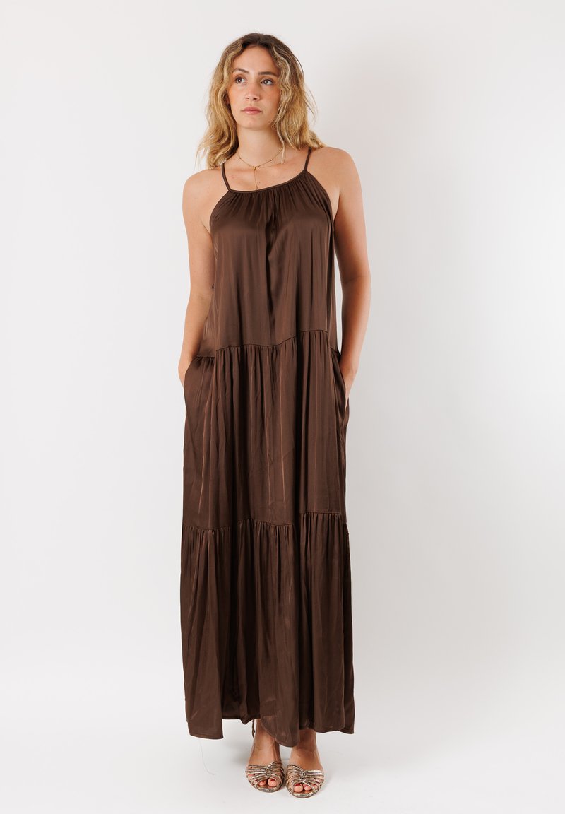 Abito maxi marrone in tessuto leggero con design a strati, presenta sottili spalline e tasche laterali. Texture liscia, silhouette fluida.