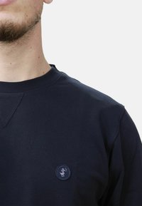 Felpa in cotone navy con collo a giro e piccolo patch rotondo con logo sul petto. Presenta un dettaglio di cucitura a triangolo all'altezza del collo.