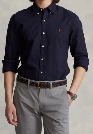 Homme portant une chemise bleu marine foncé à boutons avec les manches retroussées, un pantalon gris, une ceinture tressée marron et une montre-bracelet marron.
