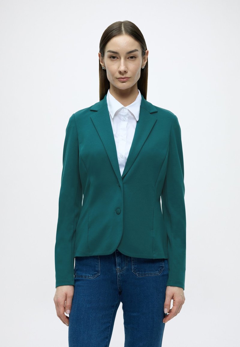 Blazer bleu sarcelle en tissu structuré, avec une fermeture à un bouton, des revers crantés et des coutures inclinées, associé à une chemise blanche à col.