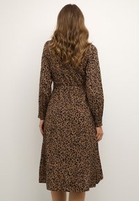 Brun midi-klänning med svarta prickar, långa ärmar och midja med bälte. Slät struktur, A-linjeform och samlade detaljer vid mässingarna.