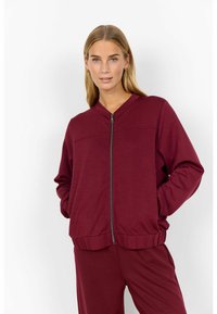 Kvinna med blont hår iförd en vinröd zip-up jacka och matchande byxor stående mot en vit bakgrund med händerna i fickorna.